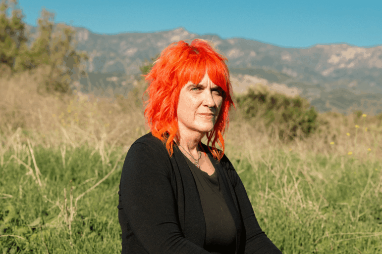 Micky Small, 53, is a screenwriter and is one of hundreds of millions of people who regularly use AI chatbots. She has bright orange, shoulder length hair and is seated with a background of mountains and brush behind her.