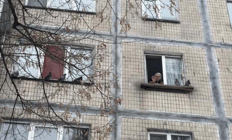 Nelia Stepanivna Thomashevska, 80, waves from her kitchen window, which is on the fourth floor of an apartment building. Two birds are perched on the windowsill.