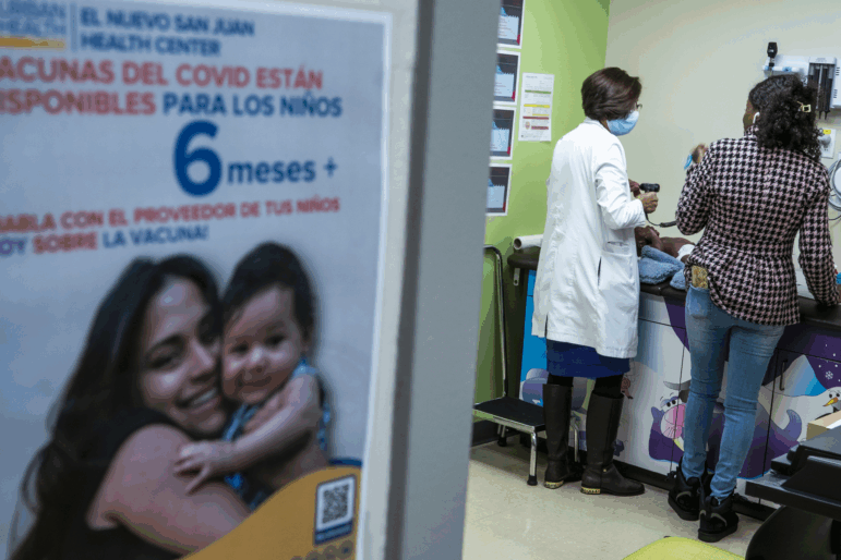 A patient care room is photographed from outside the room. A poster with Spanish language is visible, as is a doctor in a white coat, caring for a child on an exam table.