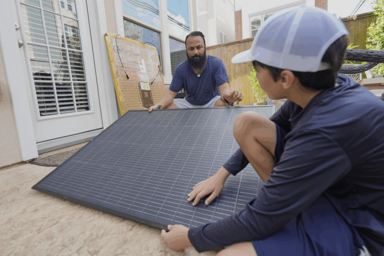 Squatting near the ground outdoors, Bhavin Misra and his son, Rumi, hold opposite sides of a black, square solar panel with a grid pattern on it while assembling a plug-in solar kit at their home in Houston.
