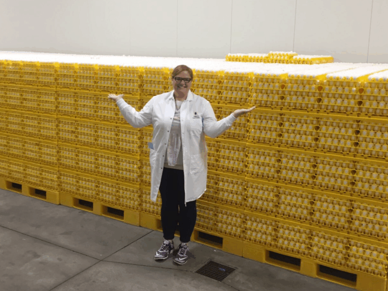 Wearing a white lab coat, dark pants and sneakers, the American Egg Board's Emily Metz stands in front of a multitude of yellow stacked trays that are filled with eggs. She is smiling and is gesturing with her arms stretched out from her sides.