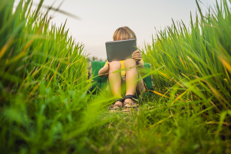 Happy child sitting on the field holding tablet. Boy sitting on the grass on sunny day. Home schooling or playing a tablet.
