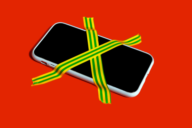 A banned smartphone is taped down to a red surface with two strips of adhesive tape that cross like an X over the phone's screen. The strips of tape have alternating yellow and green stripes on them.