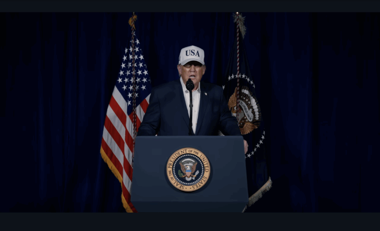 In this screenshot from a February 28 video, President Trump stands behind a lectern to discuss strikes on Iran. Speaking into a microphone, he's wearing a white baseball cap that says "USA" in navy blue capital letters on it. The presidential seal is on the front of the lectern.